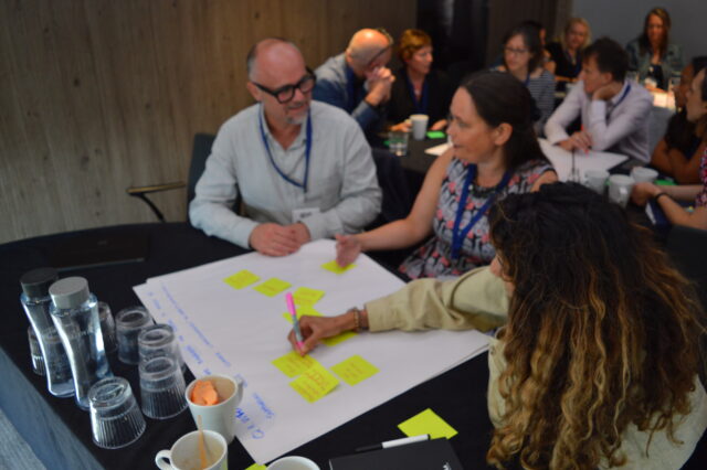 An image of three people at our collaborative workshop. They are sat around a table which has a big piece of paper on it. A woman in the foreground is writing something on a sticky note to put on the paper, and a man and a woman are sat in the background having a discussion. 