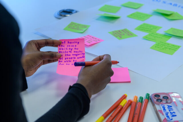 Hands holding a pink post-it note with writing on. In the background, there is a white sheet of paper with more green post-it notes on which have also been written on.