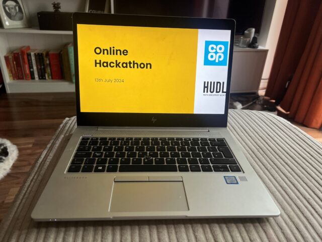 A lap top on a cream footstool that has a presentation screen up that reads: 'Online Hackathon - 13th July 2024'. Hudl creative agency and Co-op Foundation's logos also feature on the presentation slide.