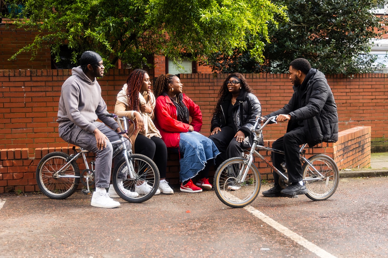 A group of young people from our funded partner, Nurturing Foundations. There are three black girls sat on a wall and two black boys sat on two bikes. They're chatting and smiling at each other.