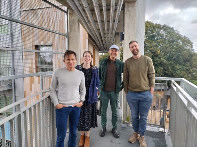 A photo of Jamie, our Chair, Ella, one of our Funding and Partnerships Managers, and representatives from RUSS, one of our partners that is a housing association we've funded with a loan. All four people are stood on a walkway in the middle of some flats. 